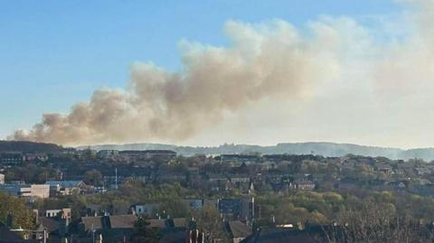 City rooftops with smoke rising up from a hill in the background against a blue sky