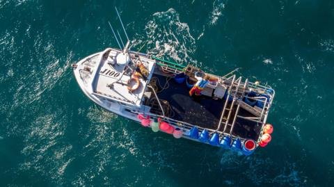 An overhead shot of a man working on a fishing boat in the North Sea. He is wearing a bright-coloured top and red trousers and is standing on the right of the boat.