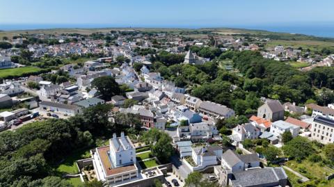 An aerial view of St Anne in Alderney. It shows rows of houses and shops form an elevated view with the sea in the background and plenty of trees and fields. 