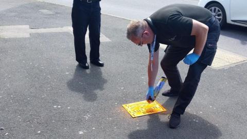 A man standing on the road wearing blue gloves and spray painting an orange mark on to the road.