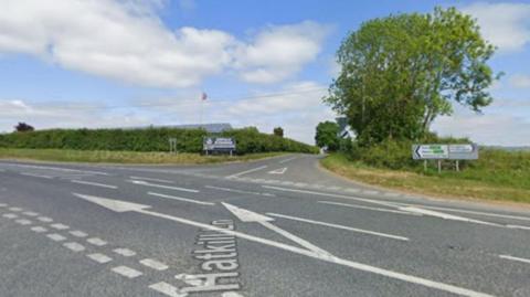 A Google maps screenshot of the junction of two rural roads, there are hedges, trees and a building in the distance.