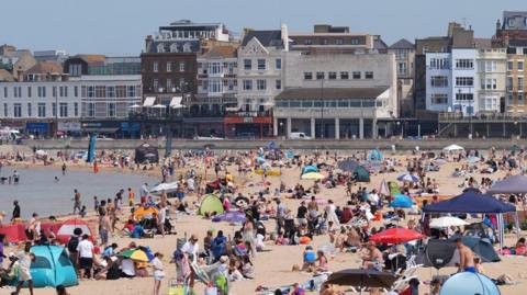 A crowded beach at Margate with large numbers of people sitting on the sand, walking along the shoreline and using tents, parasols and deckchairs. Seafront buildings and businesses can be seen in the background.