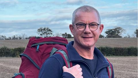 Sam Sharp standing in a field wearing a navy blue jumper and glasses. He is wearing a red backpack on his back and looking into the camera.