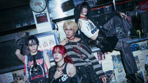 The four band members pose next to a stage in a darkly lit photo. Posters are stuck on the lower part of the stage and musical equipment is stacked behind in them in places along with a clock on a post and a digital clock below it.The performers are wearing punky-type clothing - vest tops, acidic T-shirts, jeans - and have dyed hair and jewellery to complete their androgynous look.
