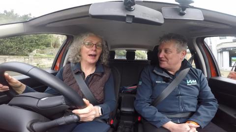 Inside a car, looking from a camera pointing towards the driver and passenger. The driver is a woman, a reporter in her 60s wearing a blue jersey shirt and brown, quilted gilet, and the passenger is a man, wearing a IAM RoadSmart zipped fleece and navy polo shirt.