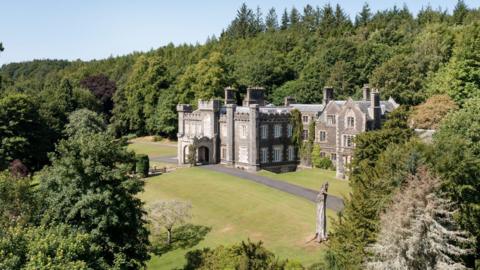 A grand stately home sits in a woodland opening. Surrounded mainly by tall green pine trees.