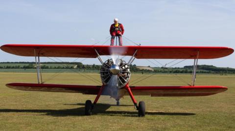 Harry is wearing a red and black jump suit and is standing on top of a red plane, in the middle of a grass landing strip at the airfield.