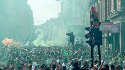 A large gathering of fans on the Trongate in Glasgow. Green and orange smoke fills the air, mostly green, and people sit up on top of traffic lights. People wear Celtic strips and wave Celtic flags 