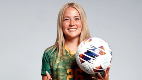 A woman with blonde straight hair standing in front of a white background. She is in a green and gold football top with the number 26 on it. She is holding a football and smiling. She has dark green shorts and long socks on, with bronze and black football boots