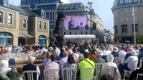 People are sat down at a previous event. They are sitting down on white chairs and looking at a large screen. There is buildings pictured around the side with a white tent at the front.