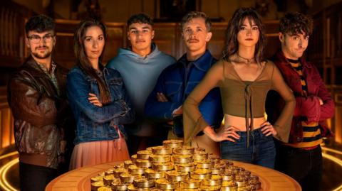 The six Traitors finalists standing behind a table with a pile of gold coins