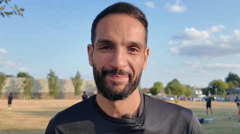 Youcef El-Barhdadi is wearing a black t-shirt and has a black beard and short dark hair. Behind him, a football training session is taking place.