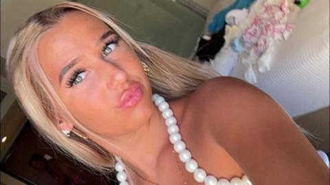 A blonde teenage girl posing for the camera. She is wearing a large necklace.