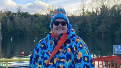 Mel Pardoe in a bright blue, yellow and orange jacket with a blue woolly hat stood in front of a lake in an old quarry