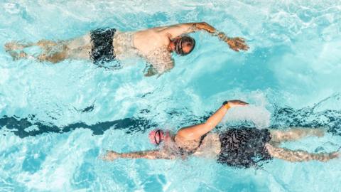 A photo taken from above of two people swimming past each other, one going to the left, one to the right of the image. They are both doing breaststroke.