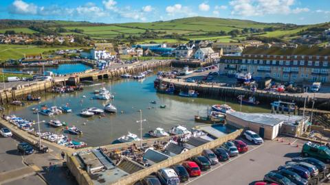 West Bay's small, square harbour has moored pleasure craft and open sides revealing fields and hills in the background.
