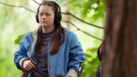 A woman wearing headphones, a light blue jacket and a stripy top underneath. Her right hand is resting on the strap of her handbag