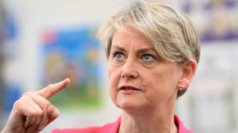 Home Secretary Yvette Cooper wearing a pink jacket looks upwards as she points a finger on her right hand to her left. She is stood in front of a wall with colourful paintings on it.