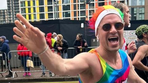 A man running a race is wearing a rainbow coloured outfit and hat, along with sunglasses. He is waving to the camera. 