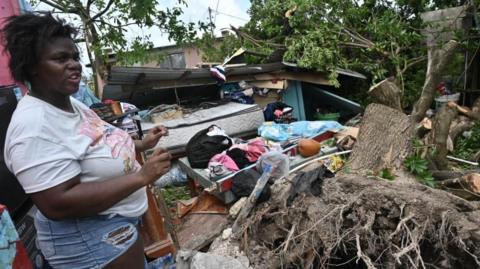 A woman stands in front of a damaged home, gesturing as she speaks. She is wearing a white T-shirt and denim shorts, behind her, fallen trees and large roots are tangled among debris from the destroyed house. Household items such as clothes, bedding and furniture are scattered amid the wreckage. The roof appears collapsed and a large tree trunk lies across the ruins.