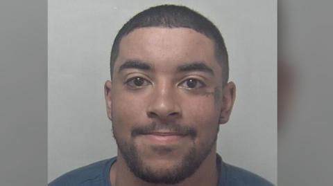 A man with brown eyes, stubble, and short cropped hair is pictured in a police mugshot. 