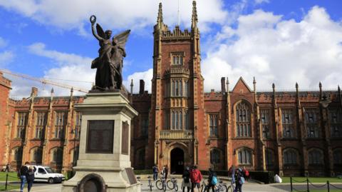 The exterior of Queen's University Belfast, a large gothic redbrick building with a statue in the font of an angel. There are some people