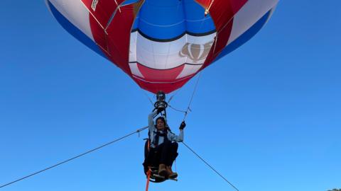 Jo Bailey is sat in a single person hot air balloon in the sky with ropes tied on either side. 