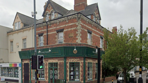 The brick exterior of Ron Greig Estates. It is a three storey brick building with green paint on the bottom floor. Properties are displayed in the windows of the ground floor. A medium sized tree is growing to the right of the building.