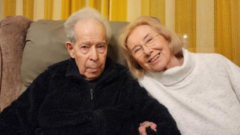 Eva and Herbert King sitting next to each other. Herbert is wearing a black top and Eva is wearing a white-grey jumper.