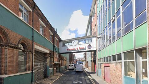 Buildings with dozens of windows line two sides of a street. A first-floor bridge in the middle of the street, which links the two buildings, has a sign which reads, "Express & Star". A number of vehicles, including vans, can be seen parked in the distance.