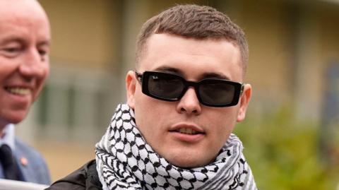 A close-up image of Liam Óg Ó hAnnaidh outside court. He is a young man wearing a pair of black RayBan sunglasses. He has a white and black keffiyah around his neck. He is looking off to the side.