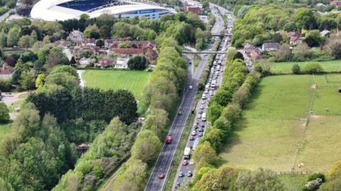 An aerial view of a busy highway which shows slow‑moving traffic queueing, with several lanes of cars and lorries backed up beside trees and a road bridge near a slip road.