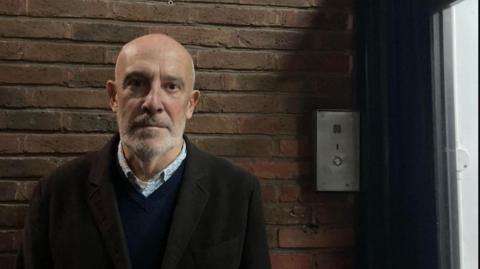 A bald man with a grey beard, wearing pale blue shirt, dark blue jumper and brown coat stares into the camera.  