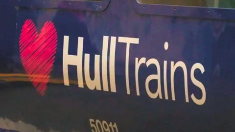 A close-up of the side of a dark blue train carriage with a large logo reading "Hull Trains" in white text, next to a pink heart. A train serial number reads "50911".