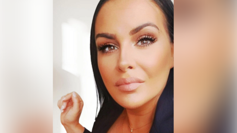 A woman with long black hair takes a selfie while standing next to a white wall.