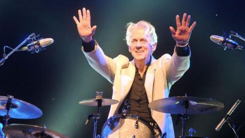 A white-haired man wearing a white suit and black shirt holds his hands in the air while sat at his drumkit. He is illuminated by stage lights and is smiling for the crowd.