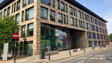 A yellow and brown brick building with large glass windows: the front door has green writing that shows the names of local places. 