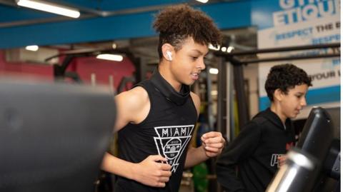 Two teenage boys at a gym - one is wearing a black vest with the word Miami in white and the other is wearing a black jumper. Both are running on treadmills.