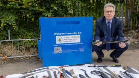 A man with greying hair and wearing a suit, crouched beside a weapon surrender bin. He is holding a large knife and there are lots of knives laid out at his feet on a blanket. 