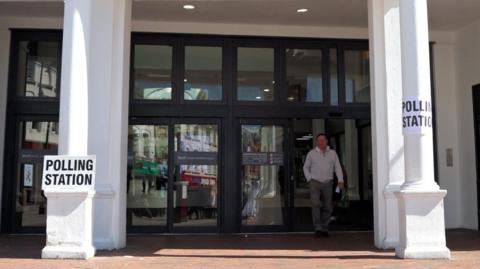 A man walking out from a large set of black doors in the front of a white building. There are two white pillars either side of the entrance with signs attached, reading 'POLLING STATION' in black lettering