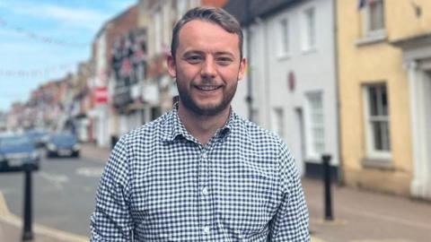 Chris Curtis stood on the high street of Stony Straford, Milton Keynes, wearing a blue check shirt. He is smiling and has short brown hair and a short brown beard.