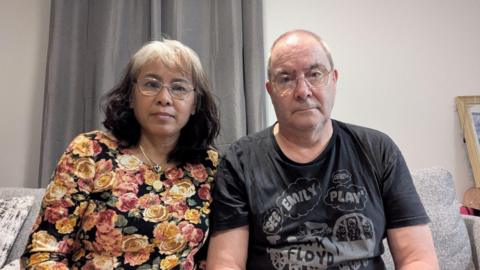 Noi Weeks and Nick Weeks, pictured at their new flat in Woking, Surrey. Noi is wearing a flowered top and has white hair and glasses. Nick is wearing a Pink Floyd t-shirt, is balding and wearing glasses.