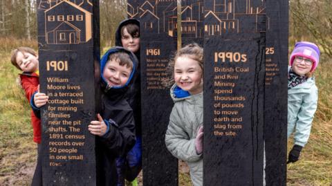 Five children stand behind black wooden planks which are installations in a woodland trail. The planks detail information about the area's mining history
