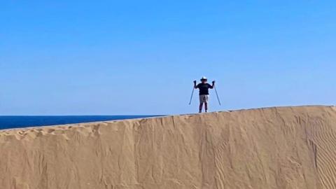 A woman in the distance trekking in sand dunes with two walking sticks wearing shorts and a hat. 