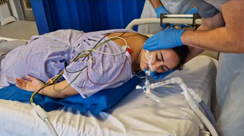 A female patient is laying on her chest, seemingly asleep, on a hospital bed. She is in a medical room with dark blue curtains separating patient beds. The patient has a medical tube attached to her nose. She is laying over a blue pillow-like device that is slowly being inflated, while a medical professional supports the patient's head.