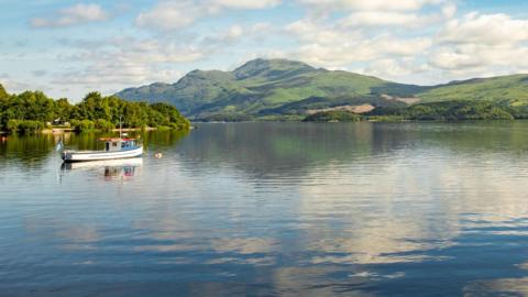 Loch Lomond, the water is blue, still and reflecting the hills in the distance. There are a few boats visible.