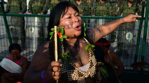 An indigenous woman protests in front of a line of soldiers. 