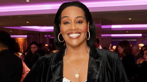 A head and shoulders shot of Alison Hammond in a black jacket posing in a room lit with purple. She has her dark hair down but combed back from her fae and wears silver hooped earrings and a heart pendant necklace. Other women can be seen chatting in groups behind her.