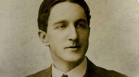 Formal portrait photo of Donnie MacRae. It is a monochrome, head and shoulders image of a young man in his 20s. He has very tidy, short, dark hair with a centre parting. He is looking slightly away from the camera with his mouth closed so that his expression is neutral. He wears a starched wing collar and tie, with a jacket over the top of his shirt. 