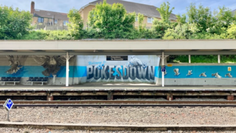 A train station platform with a blue and white mural on the back wall reading 'POKESDOWN', and railway tracks are in the foreground.